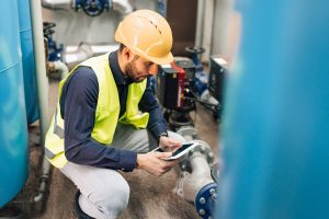 A young Caucasian male engineer is squatting and looking at his tablet, while performing a system check.